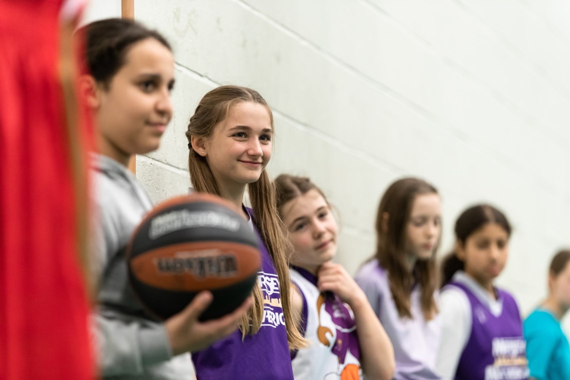 Photograph of a group of girls listening to a coach (out of shot) at a girls basketball session.